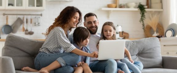 a family of four sitting close together on a couch looking at a laptop screen