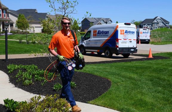 Airtron Technician Walking Into A Home With Tools In Hand And Two Airtron Service Vehicles Behind Him