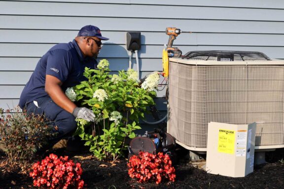 Airtron technician performing maintenance on outdoor air conditioning unit next to flowering plants