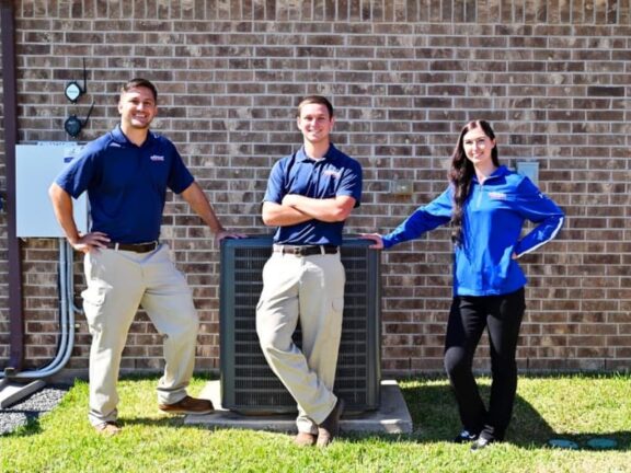 3 employees in blue shirts, outside standing in front of an air conditioning unit