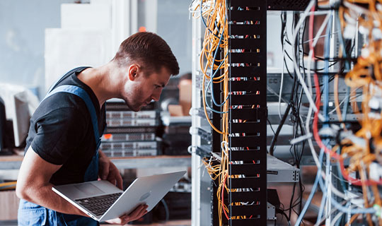 man holding laptop computer, looking at network servers in rack with wires in the foreground